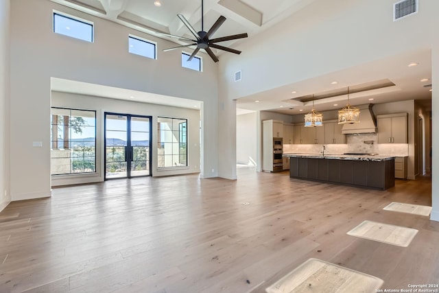 unfurnished living room with a high ceiling, coffered ceiling, ceiling fan with notable chandelier, sink, and light hardwood / wood-style floors