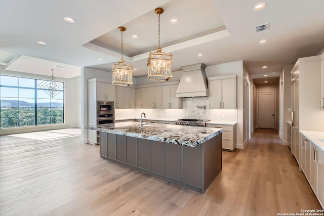 kitchen featuring premium range hood, a raised ceiling, light hardwood / wood-style flooring, light stone countertops, and a large island