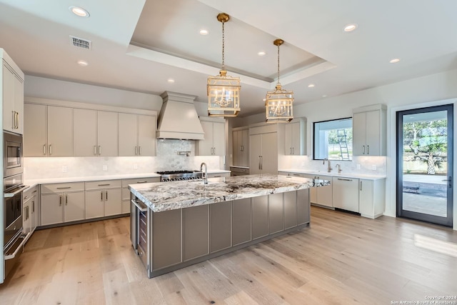 kitchen with gray cabinetry, premium range hood, a spacious island, a raised ceiling, and appliances with stainless steel finishes