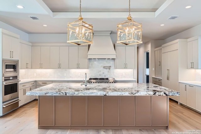 kitchen with light stone counters, custom exhaust hood, stainless steel appliances, a center island with sink, and an inviting chandelier