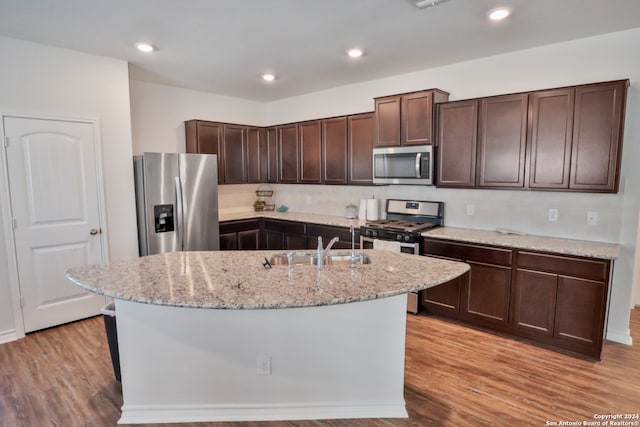 kitchen with a center island with sink, sink, light wood-type flooring, appliances with stainless steel finishes, and dark brown cabinetry