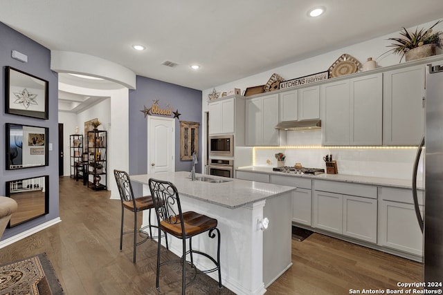 kitchen with sink, an island with sink, light stone counters, wood-type flooring, and stainless steel appliances