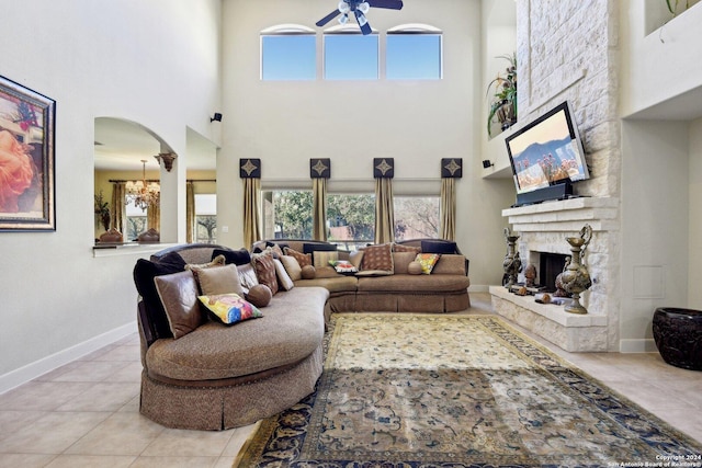 tiled living room with ceiling fan with notable chandelier, a stone fireplace, and a high ceiling