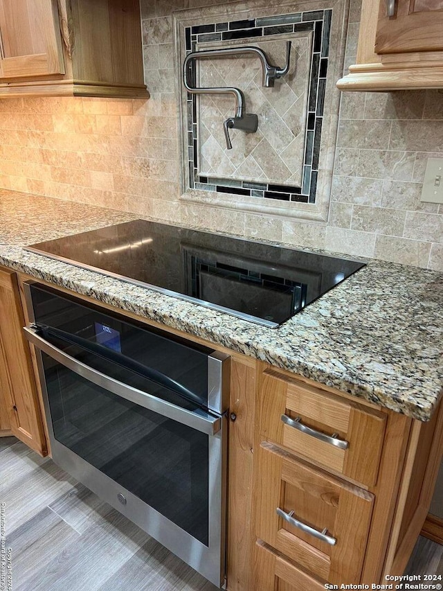 kitchen featuring black electric stovetop, decorative backsplash, brown cabinetry, light stone countertops, and oven