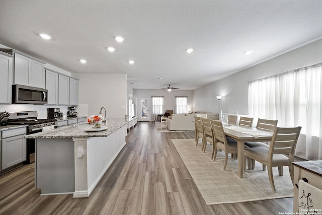 kitchen with light stone counters, gray cabinetry, stainless steel appliances, a center island with sink, and hardwood / wood-style floors