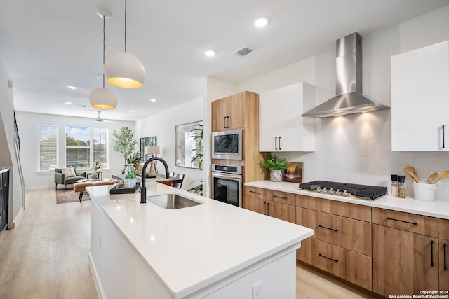 kitchen with appliances with stainless steel finishes, wall chimney exhaust hood, a kitchen island with sink, decorative light fixtures, and white cabinets