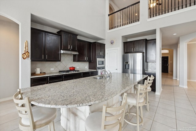 kitchen featuring a high ceiling, a kitchen breakfast bar, light stone counters, a center island with sink, and appliances with stainless steel finishes