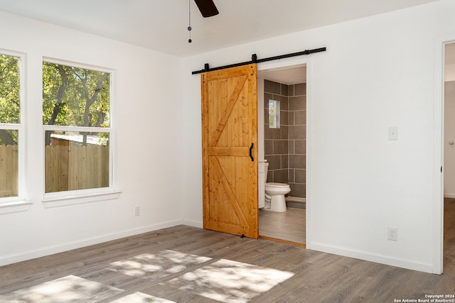 unfurnished bedroom with wood-type flooring, a barn door, ensuite bathroom, and ceiling fan