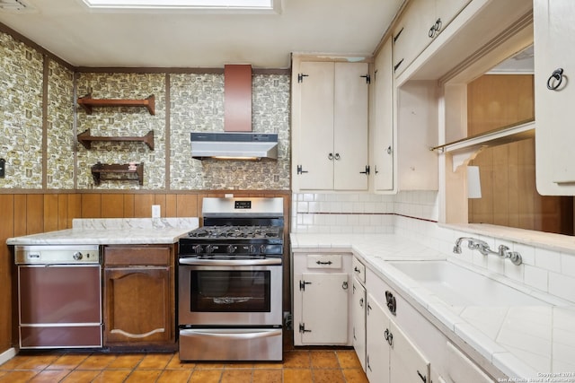 kitchen with sink, wall chimney exhaust hood, stainless steel gas range, wood walls, and white cabinets