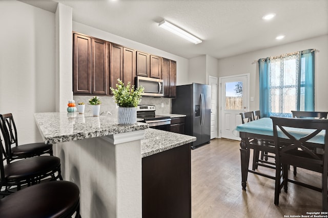 kitchen with dark brown cabinetry, light stone countertops, tasteful backsplash, appliances with stainless steel finishes, and light wood-type flooring