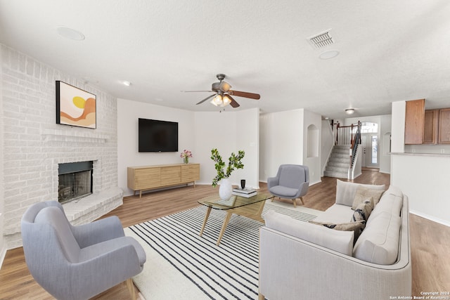 living room featuring a textured ceiling, light wood-type flooring, a brick fireplace, and ceiling fan