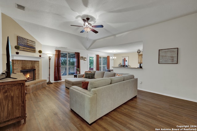 living room featuring dark wood-type flooring, ceiling fan with notable chandelier, vaulted ceiling, a brick fireplace, and a textured ceiling