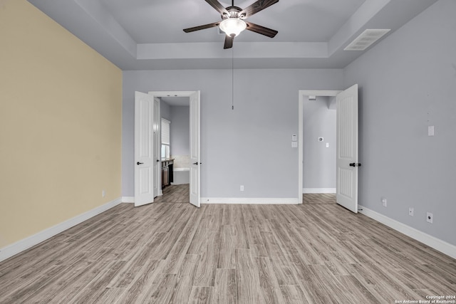 unfurnished bedroom featuring a raised ceiling, light wood-type flooring, ensuite bath, and ceiling fan