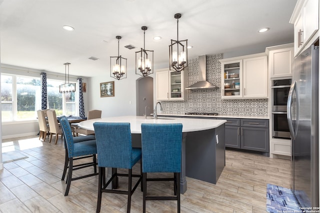 kitchen with gray cabinetry, stainless steel appliances, wall chimney range hood, pendant lighting, and a center island with sink