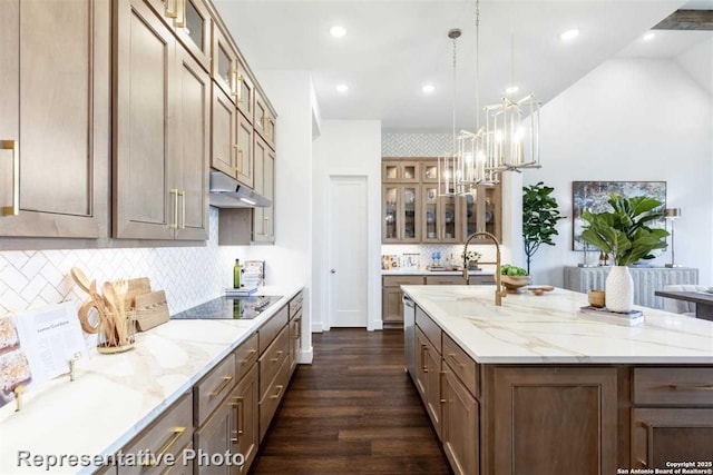 kitchen featuring pendant lighting, light stone countertops, an island with sink, black electric cooktop, and an inviting chandelier