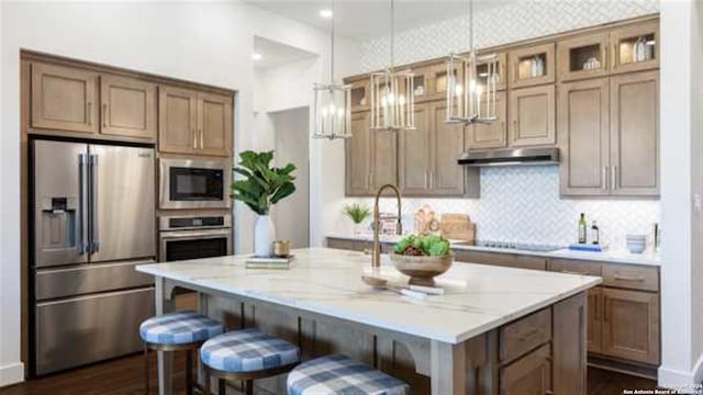 kitchen featuring a kitchen island with sink, decorative backsplash, pendant lighting, stainless steel appliances, and light stone counters