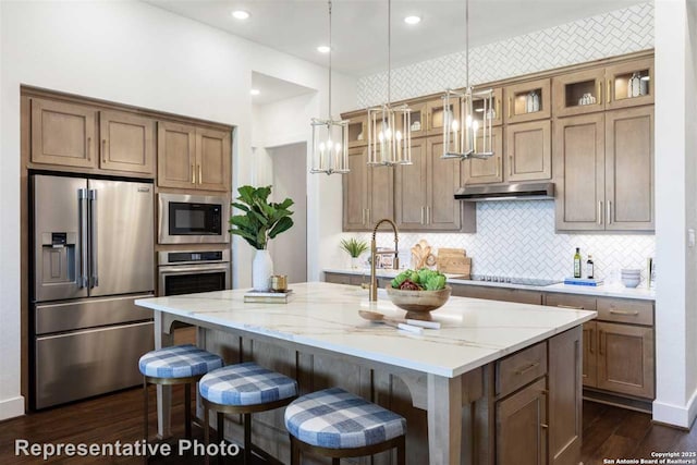 kitchen featuring appliances with stainless steel finishes, decorative light fixtures, backsplash, light stone countertops, and an island with sink