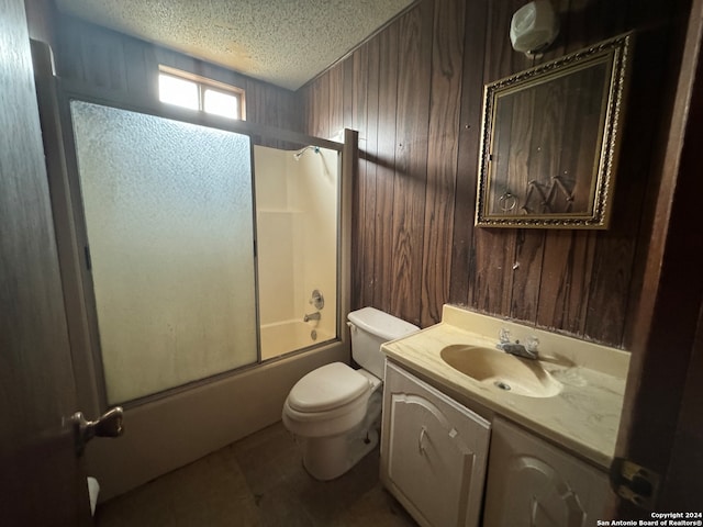 full bathroom with vanity, wood walls, bath / shower combo with glass door, toilet, and a textured ceiling