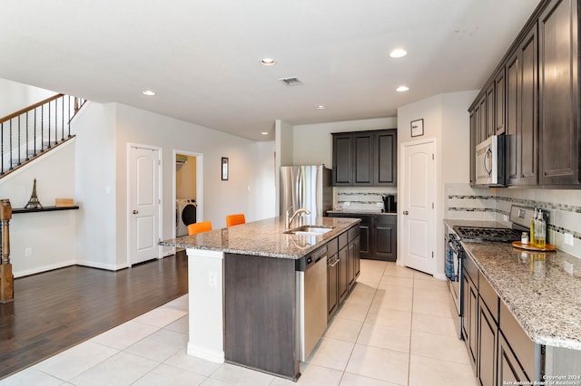 kitchen with appliances with stainless steel finishes, light wood-type flooring, light stone counters, and an island with sink