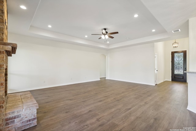 unfurnished living room with ceiling fan with notable chandelier, a tray ceiling, and dark wood-type flooring