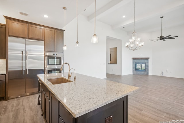 kitchen with sink, beam ceiling, stainless steel appliances, light stone counters, and an island with sink