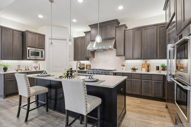 23422 Grande Vis, San Antonio, TX 78261 - kitchen featuring wall chimney range hood, dark brown cabinetry, a breakfast bar, backsplash, and light stone counters