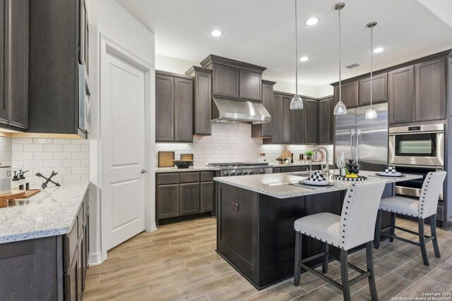 23422 Grande Vis, San Antonio, TX 78261 - kitchen with under cabinet range hood, stainless steel appliances, light wood-type flooring, dark brown cabinetry, and light stone countertops