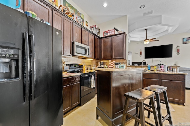 kitchen with black appliances, ceiling fan, a tray ceiling, dark brown cabinets, and a breakfast bar area