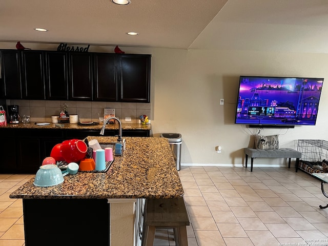 kitchen with stone counters, tasteful backsplash, light tile patterned flooring, and an island with sink