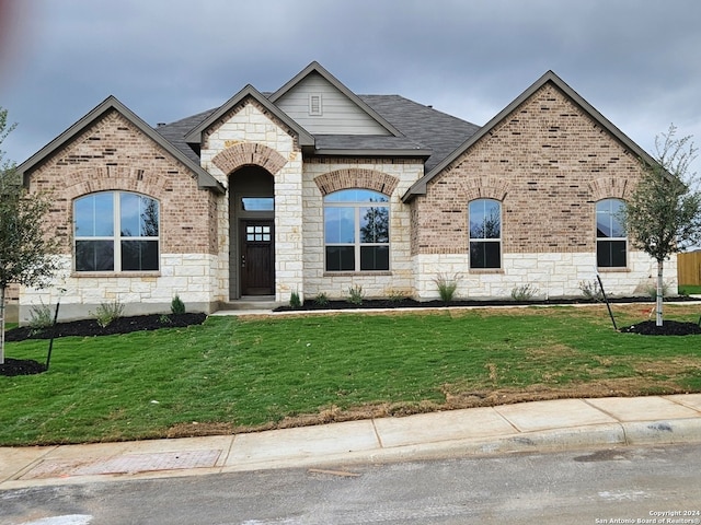 french country inspired facade with stone siding, a shingled roof, a front lawn, and brick siding