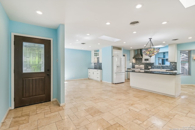 kitchen featuring backsplash, a skylight, wall chimney exhaust hood, white cabinetry, and white fridge with ice dispenser