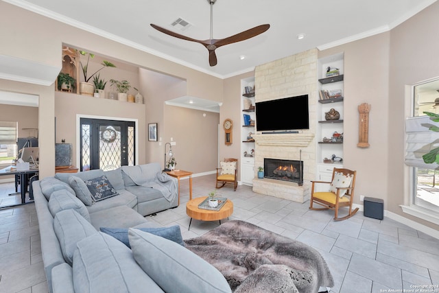 tiled living room with built in shelves, a stone fireplace, ceiling fan, and ornamental molding