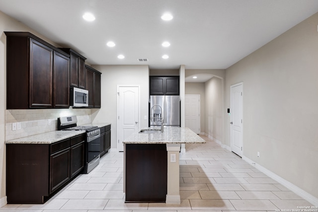 kitchen with light stone countertops, appliances with stainless steel finishes, dark brown cabinetry, a kitchen island with sink, and sink