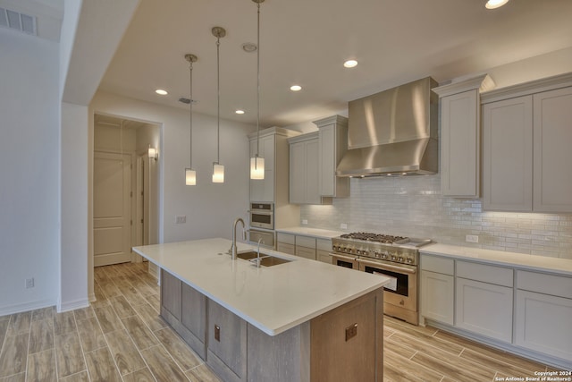 kitchen featuring appliances with stainless steel finishes, wall chimney exhaust hood, sink, a center island with sink, and hanging light fixtures