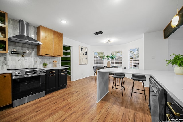 kitchen featuring stainless steel range, hanging light fixtures, wall chimney range hood, a kitchen breakfast bar, and light hardwood / wood-style floors