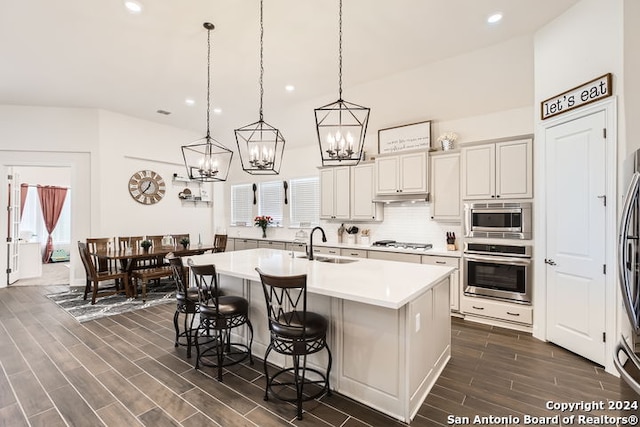 kitchen with sink, dark wood-type flooring, hanging light fixtures, stainless steel appliances, and a kitchen island with sink