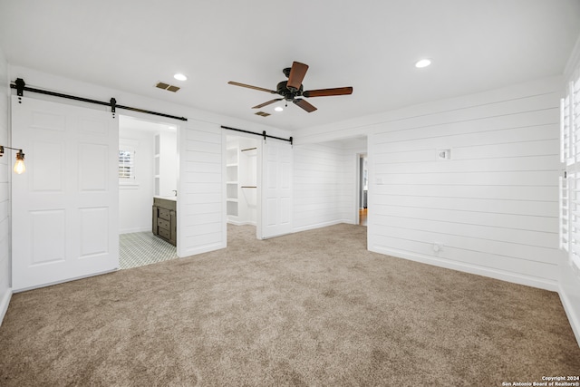 unfurnished bedroom featuring ensuite bath, ceiling fan, a spacious closet, a barn door, and light colored carpet