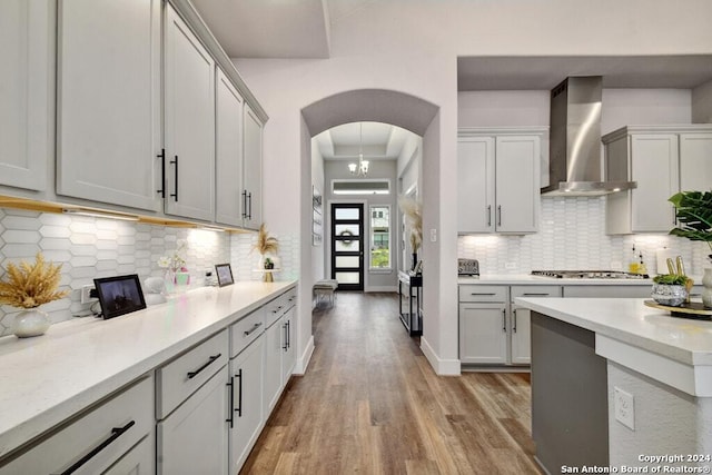 kitchen with wall chimney exhaust hood, an inviting chandelier, stainless steel gas cooktop, light hardwood / wood-style flooring, and backsplash