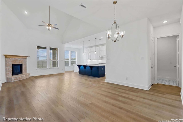 unfurnished living room with ceiling fan with notable chandelier, high vaulted ceiling, sink, a brick fireplace, and light hardwood / wood-style flooring