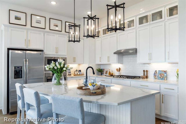 kitchen with white cabinets, an island with sink, light stone counters, stainless steel appliances, and a chandelier