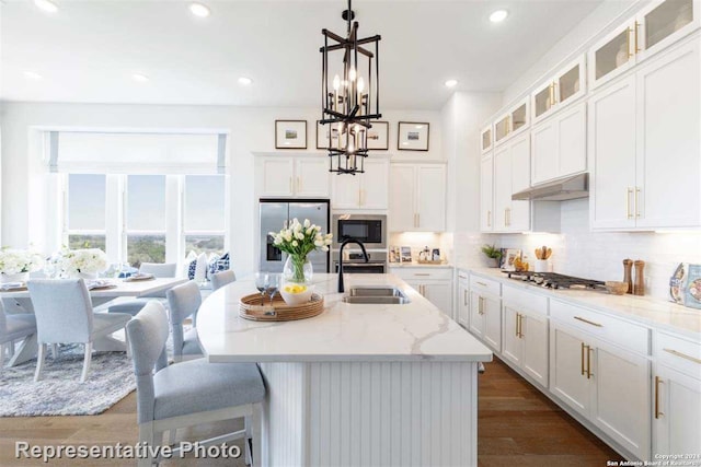 kitchen with light stone counters, stainless steel appliances, white cabinets, hanging light fixtures, and an island with sink