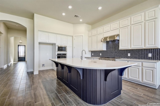 kitchen with stainless steel appliances, a kitchen island with sink, dark wood-type flooring, exhaust hood, and white cabinets