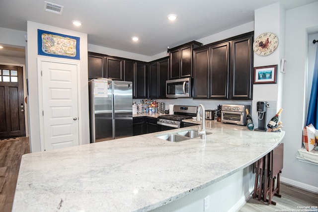 kitchen featuring wood-type flooring, kitchen peninsula, light stone countertops, appliances with stainless steel finishes, and dark brown cabinetry