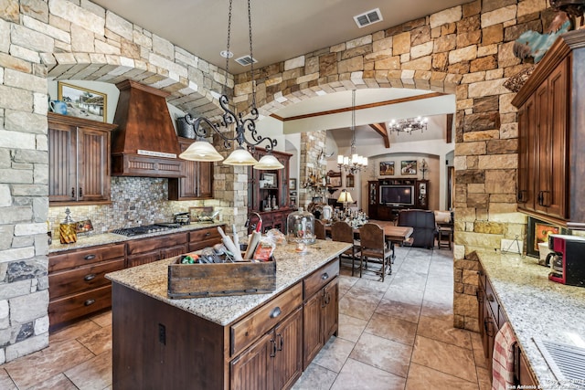 kitchen featuring light stone counters, custom range hood, hanging light fixtures, stainless steel gas stovetop, and an island with sink