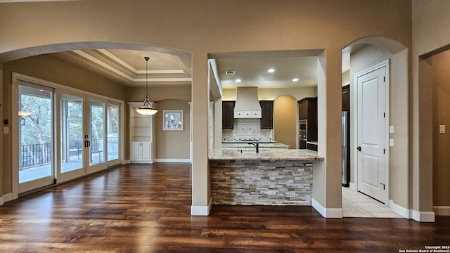 kitchen featuring a raised ceiling, wall chimney exhaust hood, ornamental molding, light stone counters, and wood finished floors