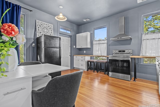 kitchen with black refrigerator, wall chimney range hood, white cabinets, light hardwood / wood-style floors, and stainless steel electric range