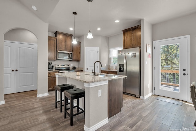 kitchen featuring light wood-type flooring, light stone counters, stainless steel appliances, a center island with sink, and hanging light fixtures