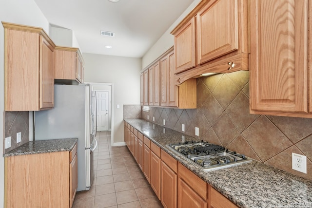 kitchen with stainless steel gas cooktop, dark stone countertops, refrigerator, decorative backsplash, and light tile patterned flooring