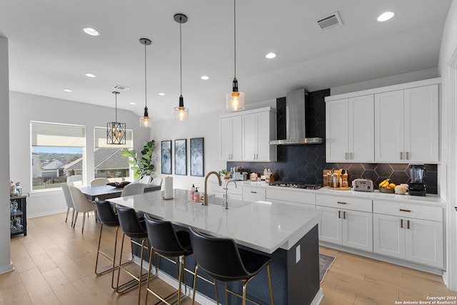 kitchen featuring wall chimney exhaust hood, an island with sink, decorative light fixtures, and light hardwood / wood-style flooring