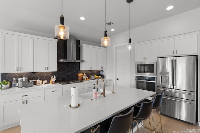 kitchen with a kitchen island with sink, light hardwood / wood-style flooring, wall chimney exhaust hood, and appliances with stainless steel finishes